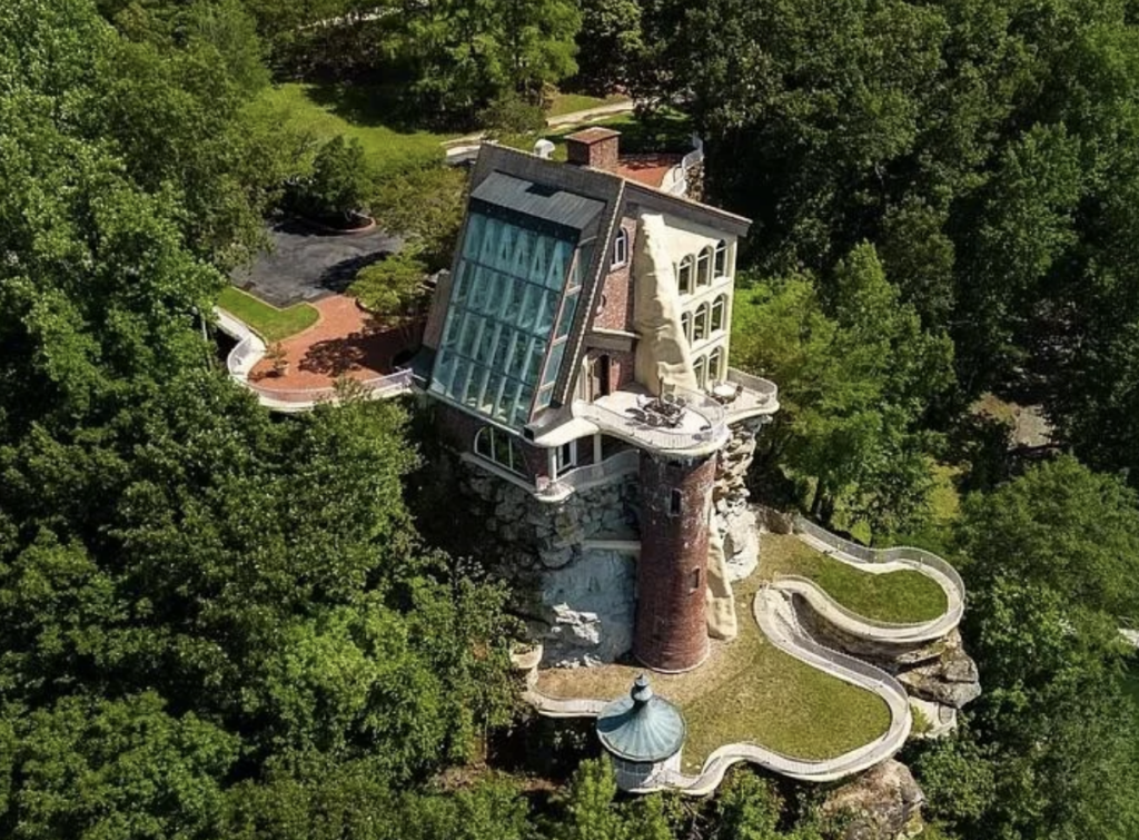 Aerial view of a unique, multi-story house built on a rocky cliff surrounded by lush greenery. The house features a mix of architectural styles, including a large glass wall, a red brick chimney, and terrace levels. A curved pathway leads to the entrance.