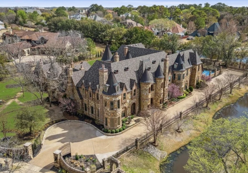 An aerial view of a grand, castle-like mansion with multiple towers and turrets, surrounded by well-manicured lawns and tall trees. The house is located in a residential neighborhood and features a long driveway and a small body of water nearby.