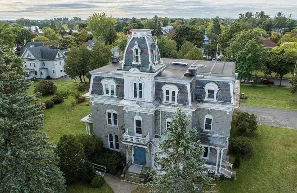 A large, historic Victorian-style house with a mansard roof and ornate architectural details stands amidst a lush green lawn. Trees surround the property, and additional houses are visible in the background under a cloudy sky.