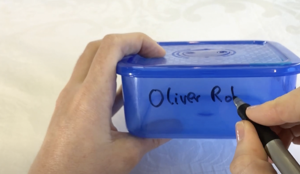 A person's hand is using a black marker to write "Oliver Rob" on the side of a small blue plastic container with a matching blue lid. The background appears to be a light-colored tablecloth.