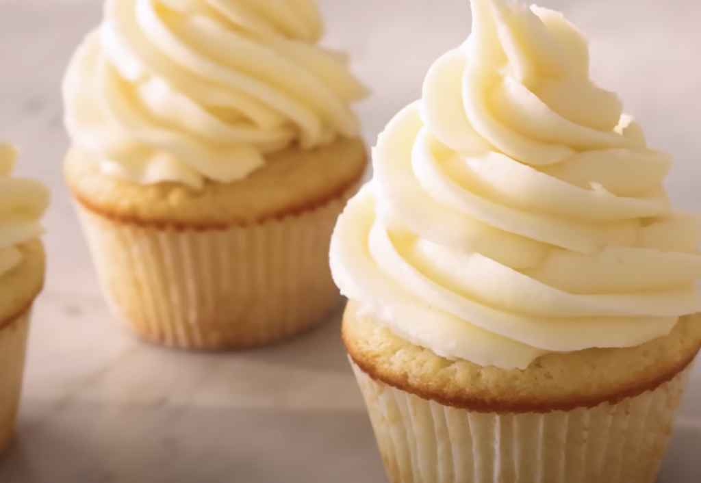 Three vanilla cupcakes with swirls of creamy, white frosting on top are arranged on a light-colored surface. The cupcakes have a moist and fluffy appearance with a generous amount of frosting.