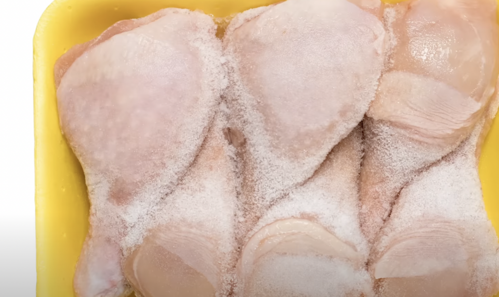 A close-up view of several frozen chicken drumsticks arranged in a yellow plastic tray. The drumsticks are covered in a thin layer of ice, indicating that they are frozen.