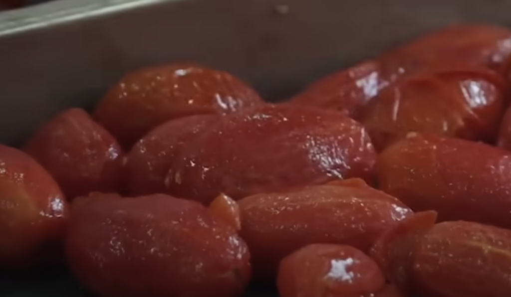 Close-up of several peeled, whole tomatoes placed together. The tomatoes have a glossy, smooth texture and are a vibrant red color, suggesting they might be freshly prepared or cooked. The background includes a faint view of a metallic surface.