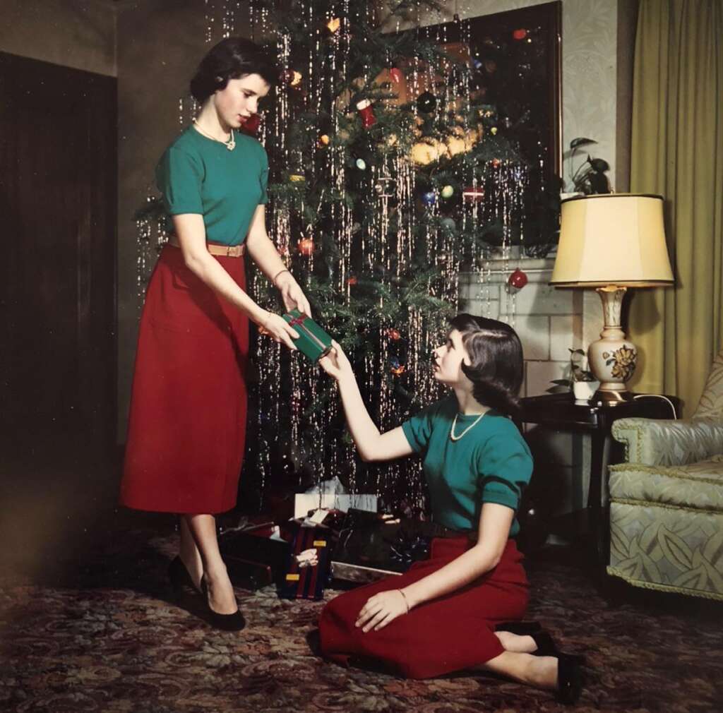 Two women dressed in matching green tops and red skirts are exchanging a gift in front of a decorated Christmas tree. One woman stands while the other kneels, both smiling. There's a lit lamp on a table and yellow curtains in the background.