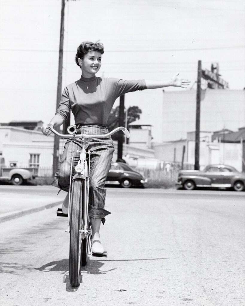 A smiling woman wearing a long-sleeve shirt and rolled-up jeans is riding a bicycle with one hand on the handlebars while extending her other arm out to the side. She appears to be cycling on a quiet urban street with a few parked cars and buildings in the background.