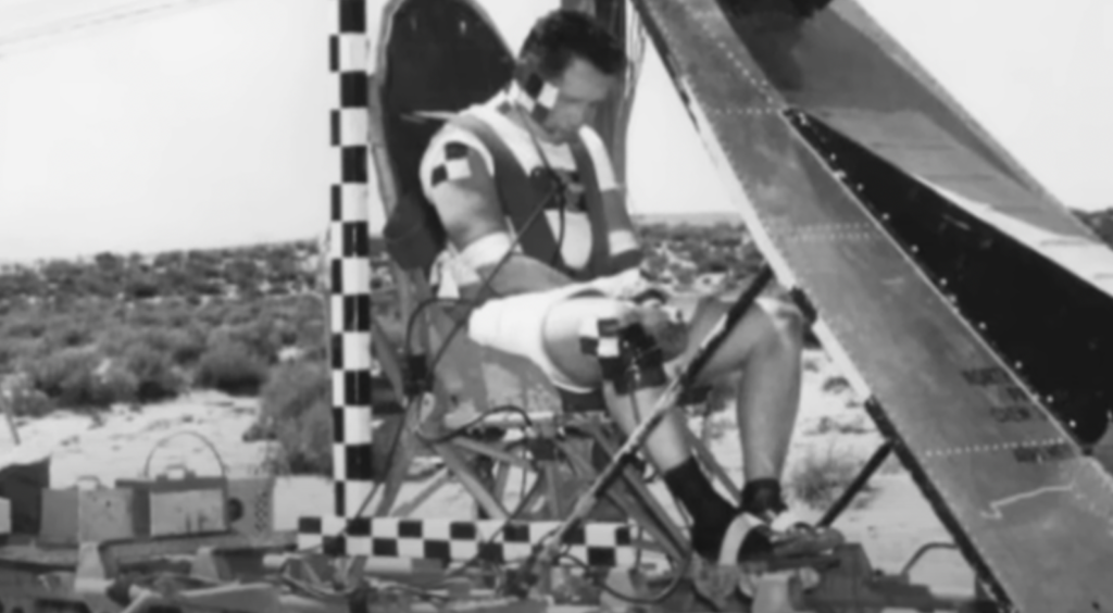 A man sits strapped to a testing machine, equipped with sensors and monitoring devices, in an open outdoor environment. The machinery features checkered patterns, and the man appears focused on an experiment or test, possibly related to aerodynamics or aviation.