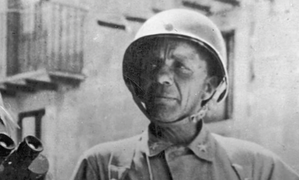 A black and white photo of an older man in a military uniform and helmet standing outdoors. The background depicts a building with balconies and a partially visible door. The man has a serious expression and appears to be holding binoculars.