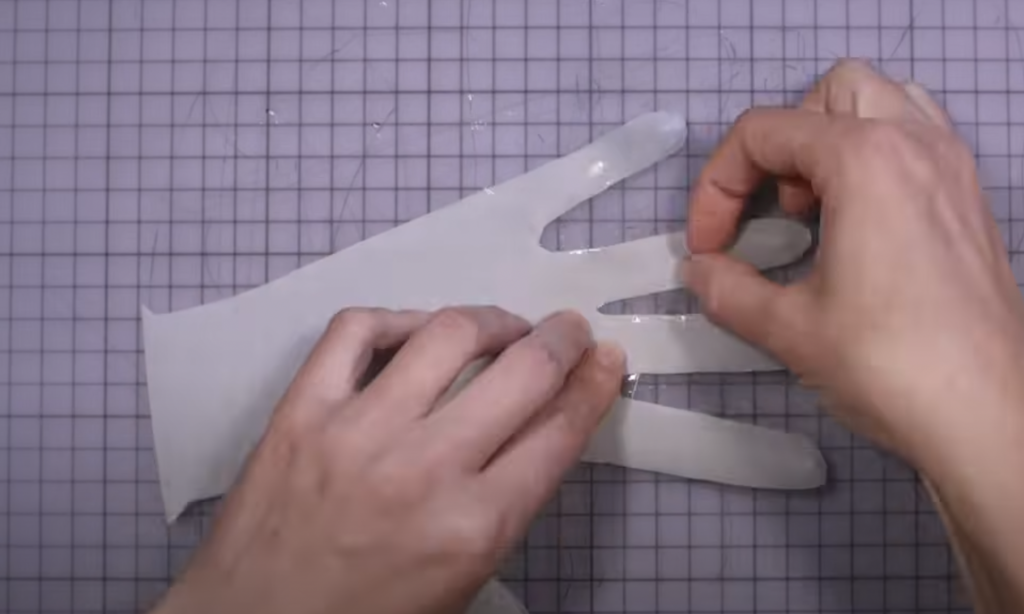 Two hands are seen in the process of creating a silicone glove mold. The glove design resembles a hand and is placed on a grid-patterned surface. The person appears to be adjusting the silicone material on one of the glove's fingers.