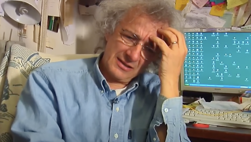 A person with curly gray hair and glasses sits in a cluttered office, looking distressed with a hand on their forehead. A messy desk with papers and a computer displaying numerous desktop icons is in the background.