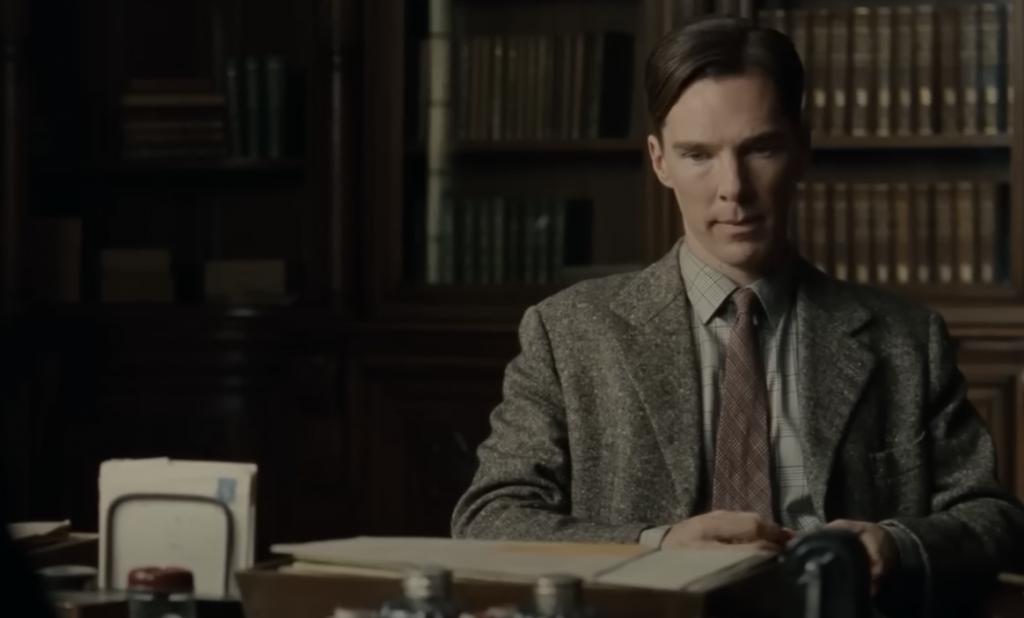 A man wearing a suit and tie is sitting at a desk cluttered with papers and office supplies. Behind him are bookshelves filled with books. He appears serious and focused while looking slightly downward.