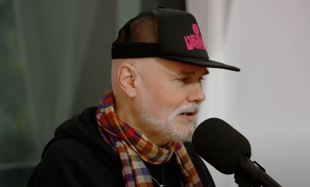 A person with a white beard is speaking into a microphone. They are wearing a black cap with pink lettering, a colorful checkered scarf, and a dark hoodie. The background is blurred, and the scene appears to be indoors.