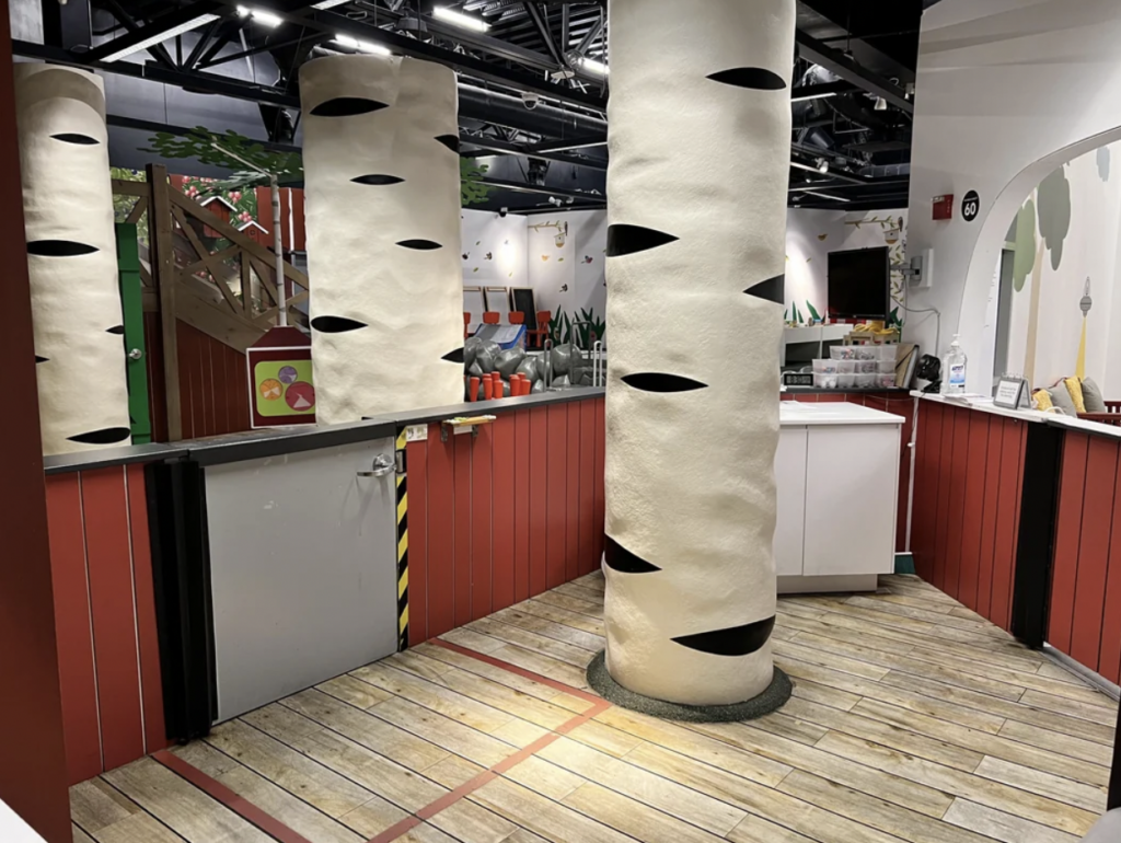 A children’s play area featuring tree-shaped columns, wooden floors, and a play structure in the background. The walls are decorated with whimsical, nature-themed artwork. A small gate and a countertop with various toys and supplies are visible.