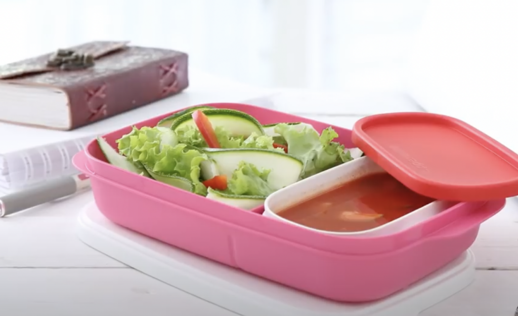 A pink bento box on a white table contains fresh salad with lettuce, cucumber, and tomatoes, and a separate section filled with soup. A red lid rests beside the box. In the background, there is a closed book and a pen.