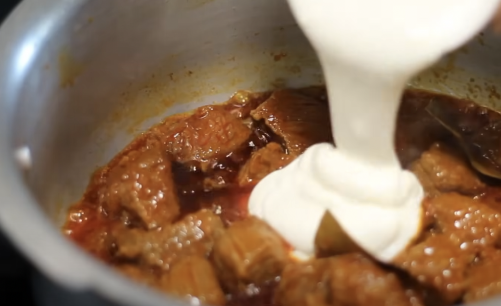 A hand is pouring a thick, creamy white substance into a pot of cooked meat with sauce. The pot is partially filled with chunks of meat in a rich, brown-colored gravy. The scene suggests the preparation process of a savory dish.