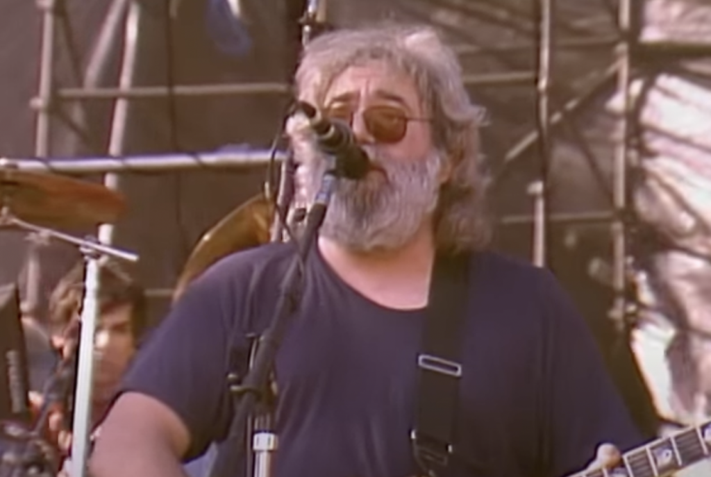 A musician with gray hair, a beard, and sunglasses sings into a microphone while playing an electric guitar on stage. There is a drum set visible in the background.