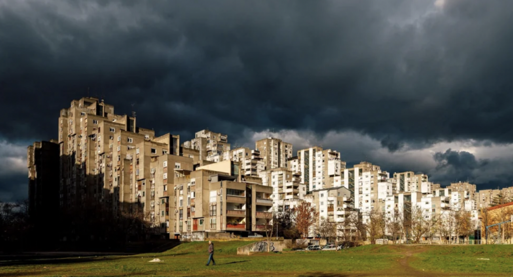 Image of a moody sky with dark clouds hanging over a large, brutalist apartment complex. The sun casts dramatic light and shadows on the buildings. A solitary person walks in the foreground on a grassy area, creating a stark contrast with the ominous sky.