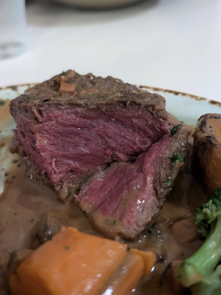 A close-up of a medium-rare steak served on a plate with creamy sauce. The steak has a pink center and is accompanied by a piece of broccoli and a slice of sweet potato. The dish is presented on a light-colored plate.