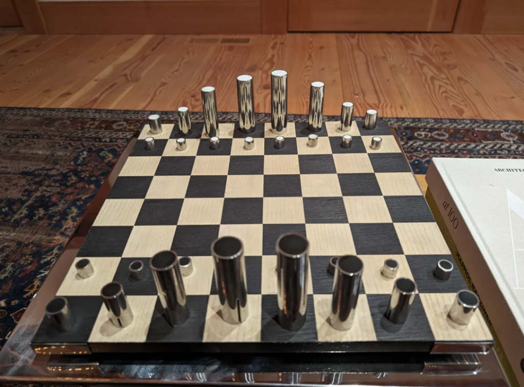 A modern metal chess set displayed on a checkered board with sleek, cylindrical pieces in varying heights. The board is placed on a wooden surface, next to a book. The background features a wooden floor and a part of a patterned rug.