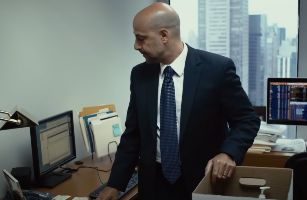 A man in a suit stands in an office, looking to his right while holding a box. Two computer monitors, various folders, and office supplies are on the desk. Through the window, a cityscape is visible.