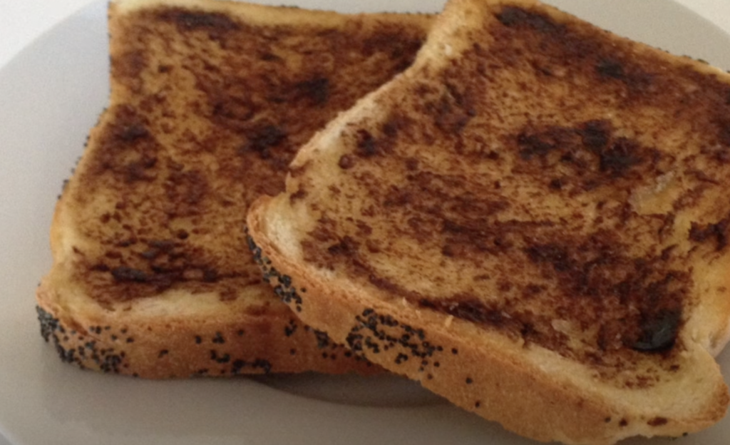 Two slices of toasted bread sit on a white plate. The toast is browned and has a slightly uneven texture with some darker, more toasted spots. The crust edges have a sprinkling of poppy seeds.