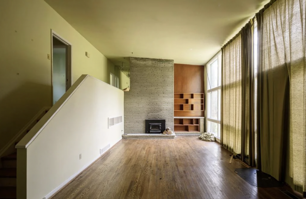 The image shows an empty living room with wooden floors and a slanted ceiling. The far wall has a tall, narrow brick fireplace, flanked by built-in wooden shelves on the right. Two large windows with floor-length curtains are on the right side, and a staircase is visible to the left.