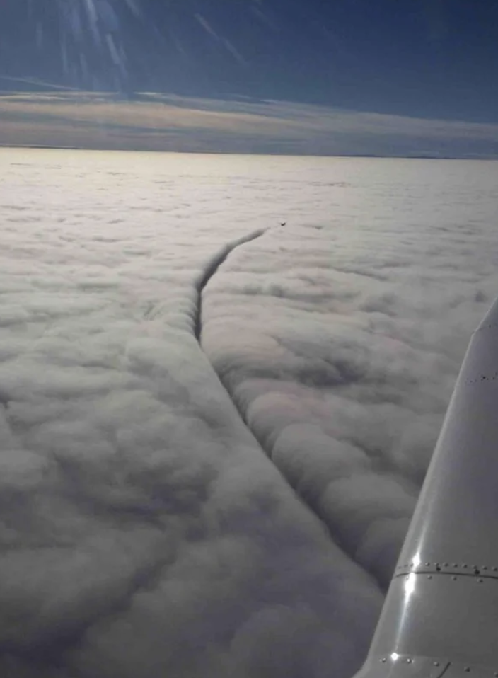 View from an airplane showing a vast expanse of clouds with a long, narrow, and straight line cutting through the cloud field. The airplane's wing and a part of the engine are visible on the right side of the image. The horizon is clear with a blue sky.