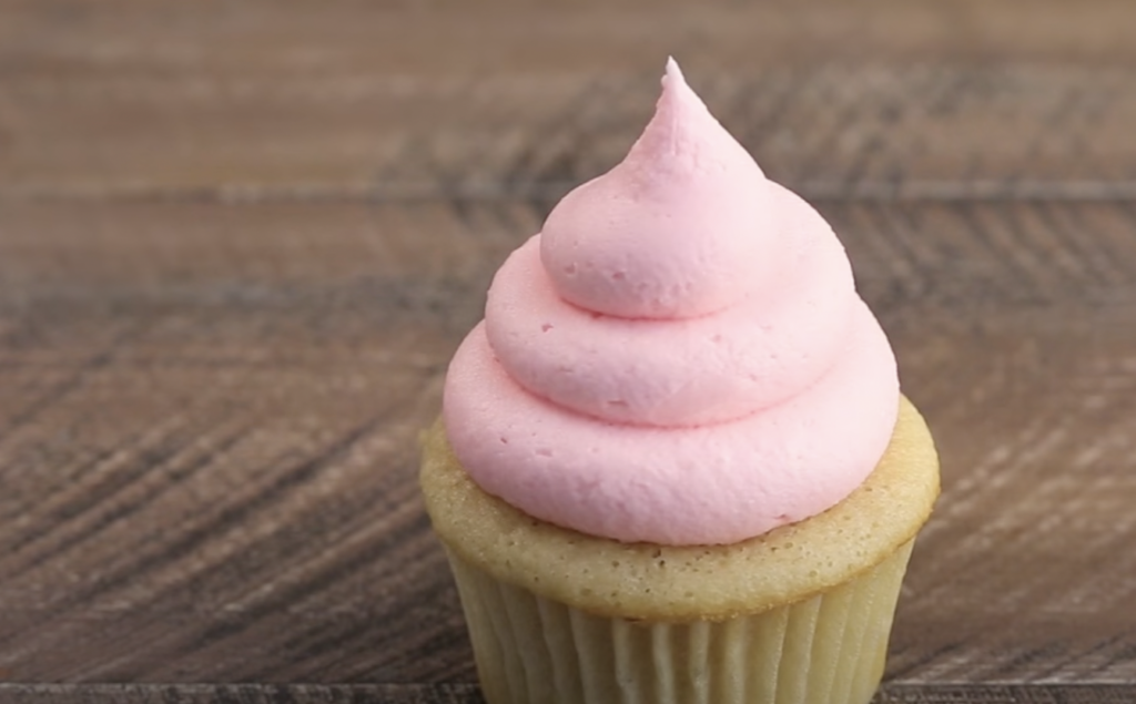 A vanilla cupcake with light pink frosting swirled in a tall, pointed peak sits on a wooden surface. The frosting is smooth and creamy, contrasting with the rustic texture of the wooden background.