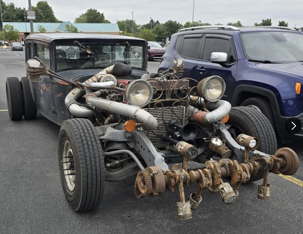 A rusty, heavily modified car with exposed engine parts, large pipes, and unique, unconventional design features, parked next to a blue SUV in a parking lot. Trees and buildings are visible in the background.