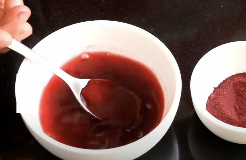 A person stirring a dark red liquid in a white bowl with a metal spoon. Another white bowl filled with a reddish powder is next to it, placed on a black countertop.