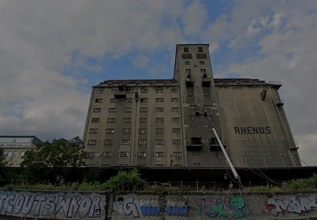 A large, weathered, concrete building with the word "RHENUS" on it stands against a cloudy sky. Below is a row of colorful graffiti on a wall, partially hidden by greenery. Scaffolding and cranes are visible, indicating construction or renovation work.