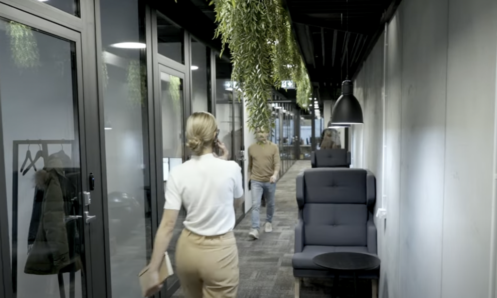 A modern office hallway with glass-walled rooms on the left. A person is walking towards the right, holding a phone to their ear. Another person is at the far end of the hallway. Potted plants hang from the ceiling, with seating and a small table on the right side.