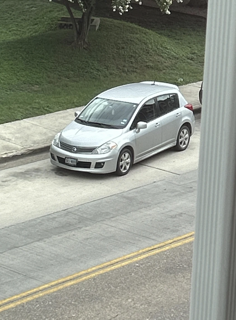 A silver compact sedan is parked on a residential street near a grass hill. The car is facing to the left and positioned parallel to the sidewalk. There is a yellow line marking on the road, indicating a two-way street.