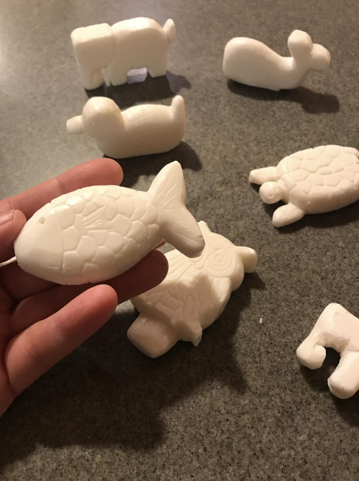 A hand holding a white, fish-shaped soap bar. Several other animal-shaped soaps, including a turtle, an owl, a dog, and a dolphin, are arranged on a gray countertop in the background.