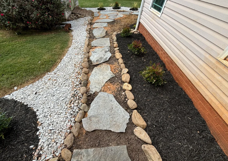 A garden pathway with large irregular stone slabs bordered by small rocks winds alongside a house. The garden on one side has white pebbles, while the other side features dark mulch with small shrubs. A grassy lawn and a flowering bush are in the background.