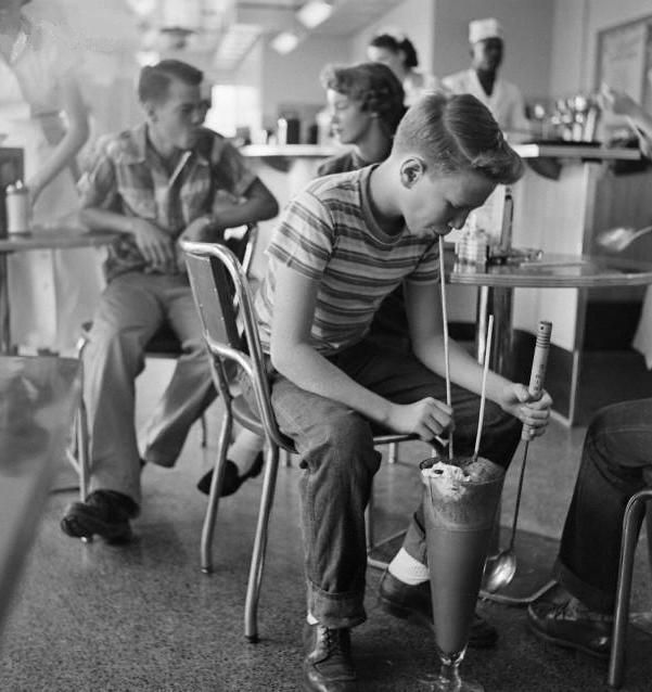 A black-and-white photo of a boy in a striped shirt sitting at a diner and drinking a large milkshake with a long straw. Two other teens, a boy and a girl, sit behind him at a table, and a waiter is in the background. The scene appears to be from the 1950s.