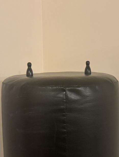 A large black cylindrical object with a textured surface is positioned against a beige wall. On top of the cylinder are two small black figurines shaped like penguins. The figures are spaced apart and facing outward.