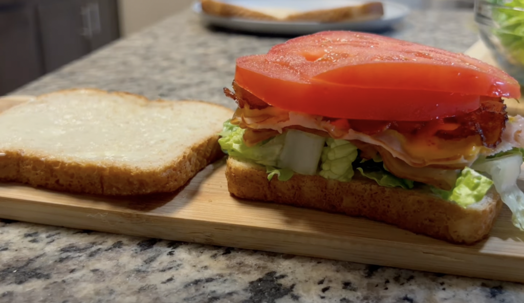 A close-up of a partially assembled sandwich on a wooden cutting board. The sandwich consists of layers of lettuce, sliced cucumber, turkey or chicken, bacon, and a thick slice of tomato. A slice of plain bread is beside the sandwich. A plate with another slice of bread is blurred in the background.