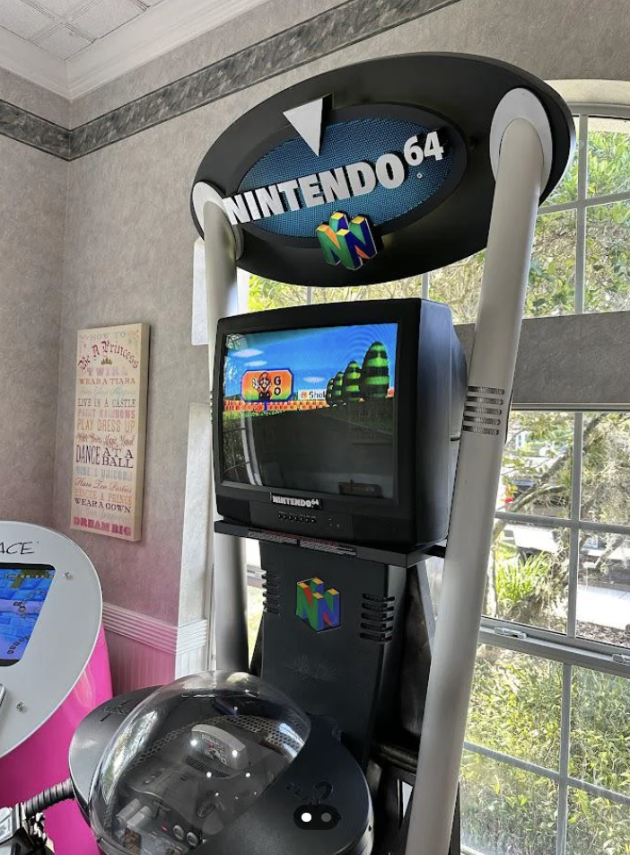 A Nintendo 64 gaming kiosk with a CRT monitor displaying a game. The kiosk features the Nintendo 64 logo prominently at the top. It stands next to a window with sunlight streaming in, and there's another machine partially visible on the left.