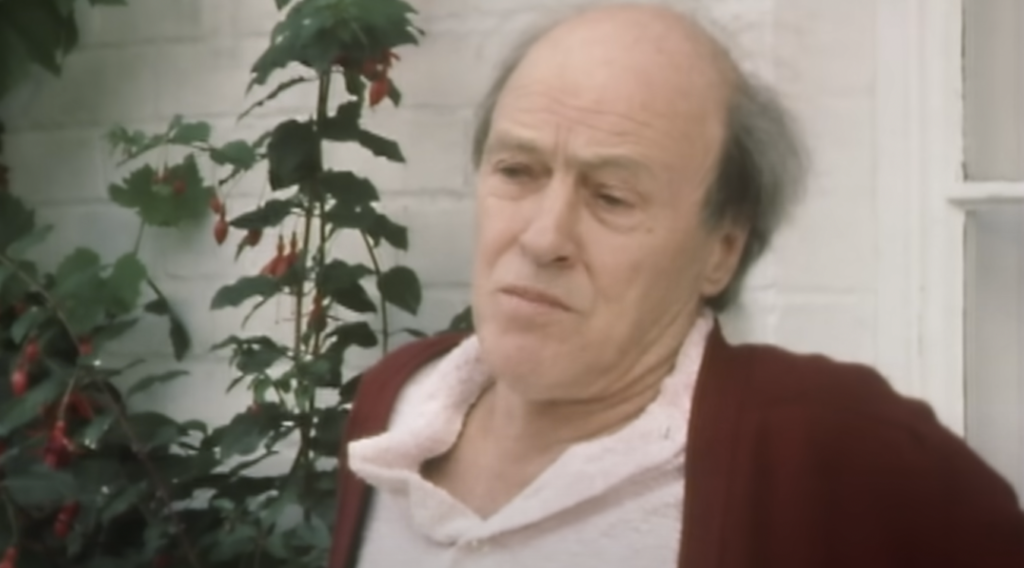 An elderly man with a receding hairline, wearing a red cardigan and white shirt, stands outside in front of a white brick wall with a flowering plant beside him. He has a thoughtful expression on his face.