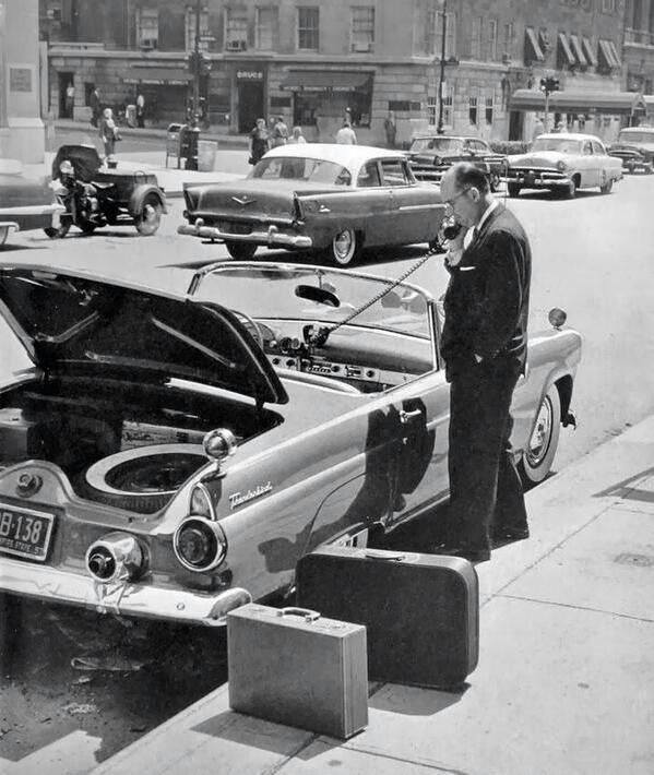 A man in a suit uses a car phone beside a classic open-top Thunderbird car with its trunk open, parked on a city street. Luggage is on the sidewalk nearby, and vintage cars and motorcycles are visible in the background. The scene appears to be from the mid-20th century.