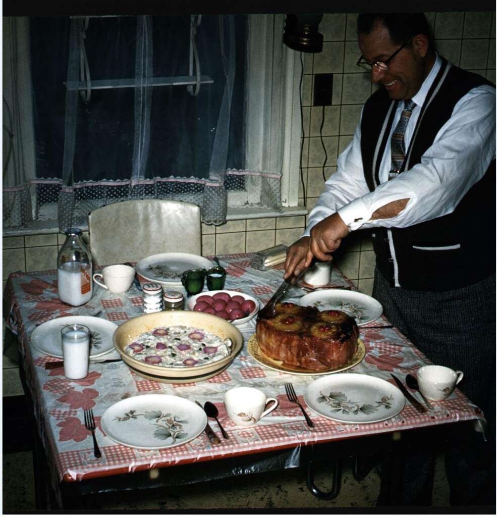 A man wearing glasses and a tie is cutting a pineapple upside-down cake on a table set with floral-patterned dishes. The table also has a salad, pickles, bright red eggs, and a milk bottle, with a checkered tablecloth and floral-patterned placemats.