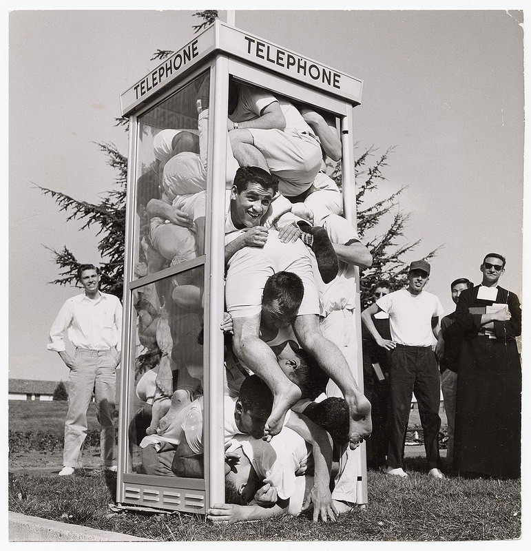 A group of young men, mostly wearing white shirts and shorts, are humorously crammed inside a telephone booth. Some are leaning out, while others seem tightly packed. A few onlookers, including a man in a black robe, stand nearby smiling and watching.