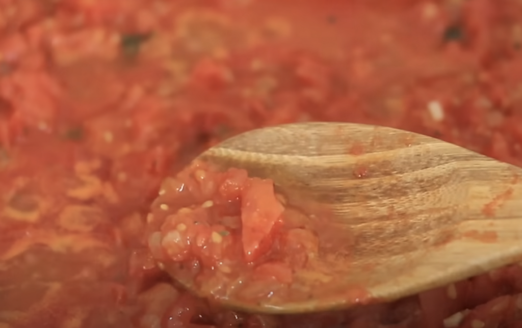 Close-up of a wooden spoon scooping a chunky, red tomato sauce that is simmering. The sauce contains visible bits of tomatoes, seeds, and other small vegetable pieces. The background shows the rest of the sauce in the cooking pot.