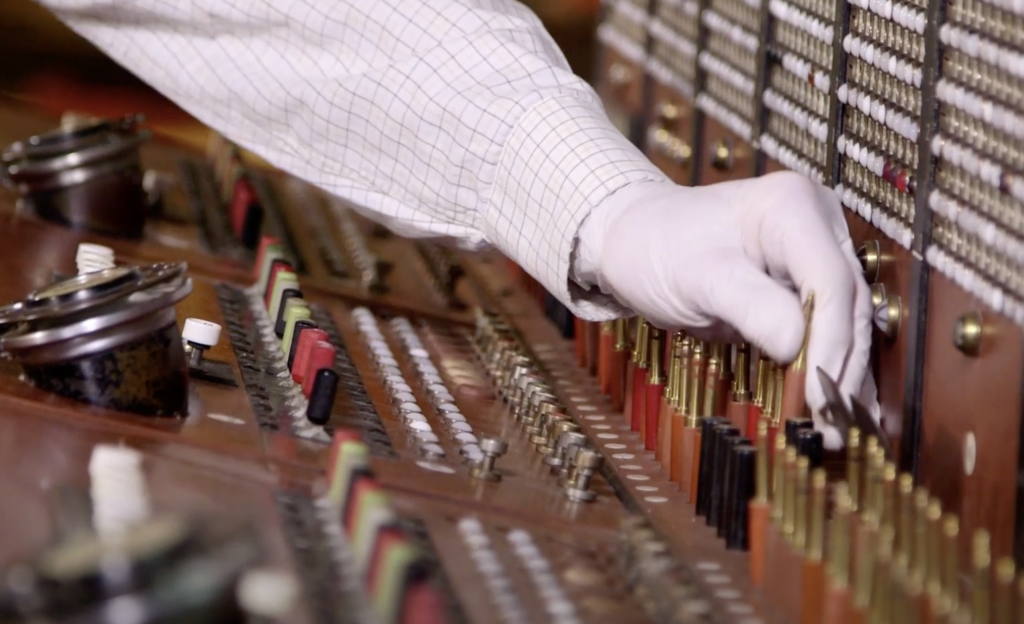 A person wearing a white glove is adjusting a control on an old-fashioned switchboard with numerous buttons, knobs, and colored wires. The background is filled with various electronic components and connectors.