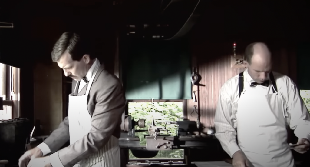 Two men wearing aprons work in a dimly lit workshop. One man, with a mustache, stands near a window examining papers. The other man, who is bald, works at a table on the right. Various tools and machinery are visible in the background.