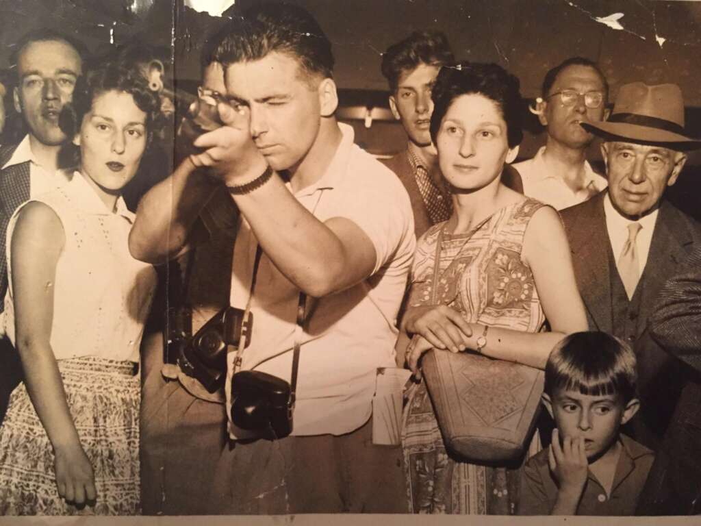 A vintage black-and-white photograph showing a group of people standing closely together. A man in the foreground holds a rifle, aiming into the distance, while a woman stands beside him. Other individuals of varying ages stand around them, observing intently.