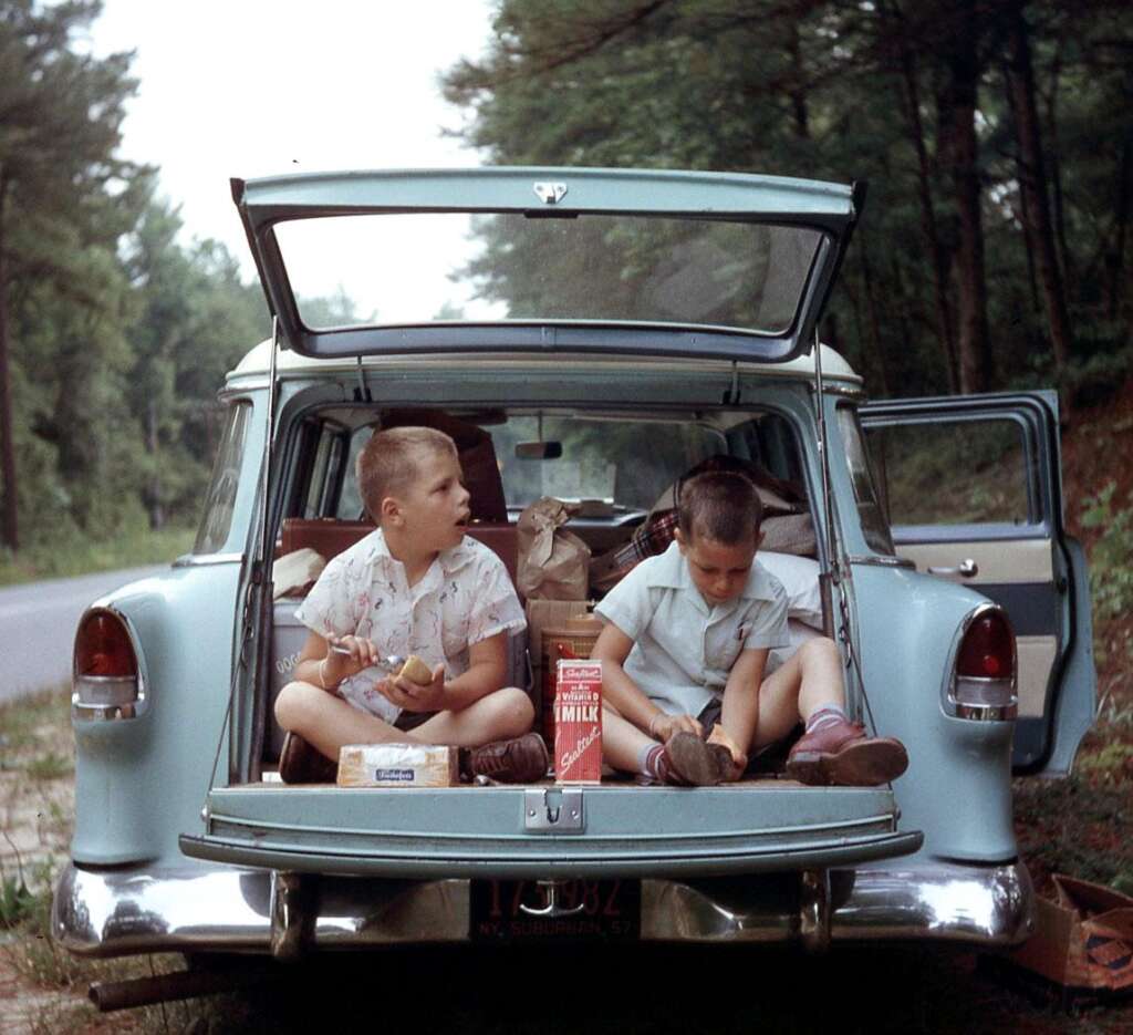 Two children sit in the open trunk of a vintage car, eating food. The trunk is filled with bags and groceries, including a visible carton of milk. The car is parked by a wooded area, with trees and a roadside in the background. The children are wearing casual clothes.