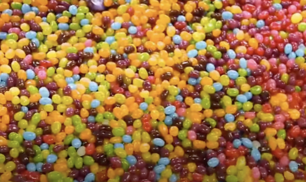 A close-up view of a large pile of colorful jelly beans. The jelly beans are mixed in various vibrant colors including red, yellow, orange, green, blue, and purple, creating a lively and cheerful appearance.