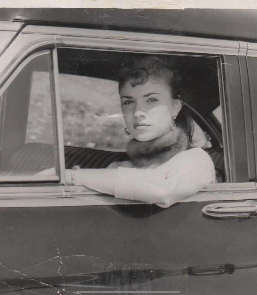 A black-and-white photograph of a woman sitting in the driver's seat with her arm resting on the open window of a vintage car. She is looking into the distance, wears a light-colored coat, and has curled hair and earrings. The car door and part of the interior are visible.