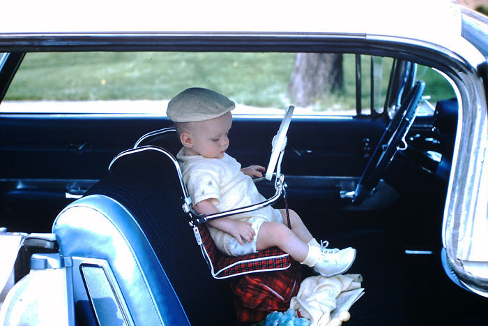 A baby, dressed in a light-colored outfit and hat, sits in a plaid-trimmed baby seat in the driver's seat of a vintage car. The car door is open, revealing a grassy area and tree trunk outside.